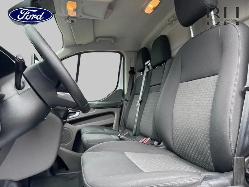 Vue intérieure côté conducteur du Ford Transit Custom blanc glacier 2023, montrant les sièges avant en tissu gris et le volant cuir.