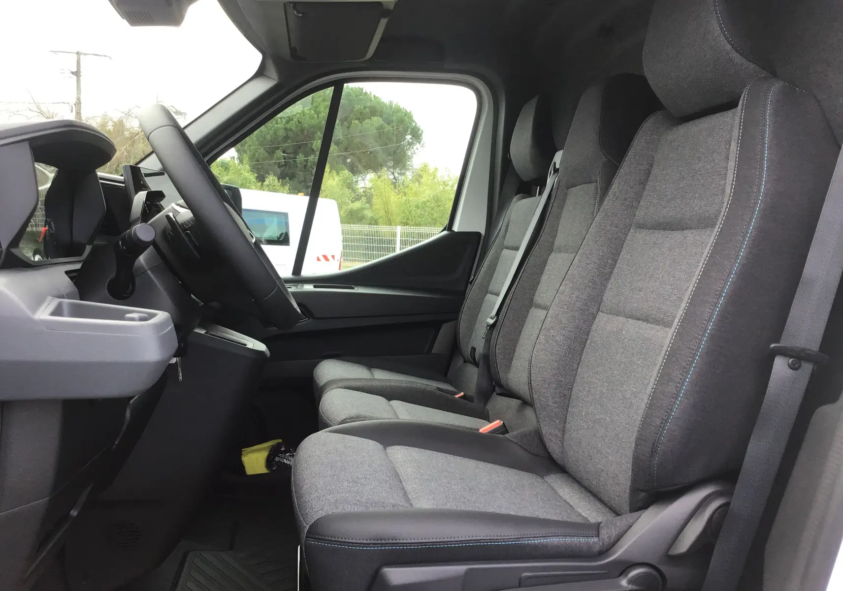 Vue intérieure côté conducteur du Renault Master blanc 2025, montrant les sièges gris avec surpiqûres bleues et le volant.
