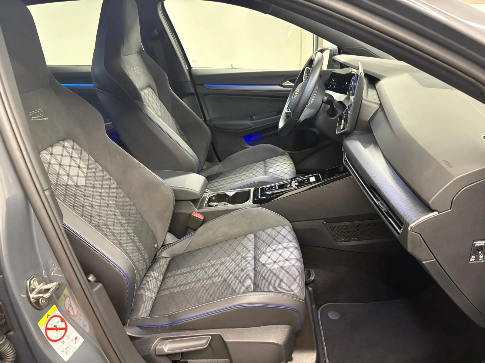 Vue intérieure côté conducteur de la Volkswagen Golf 1.5 eTSI R-Line Edition 2025 avec sièges gris à surpiqûres bleues et tableau de bord moderne.