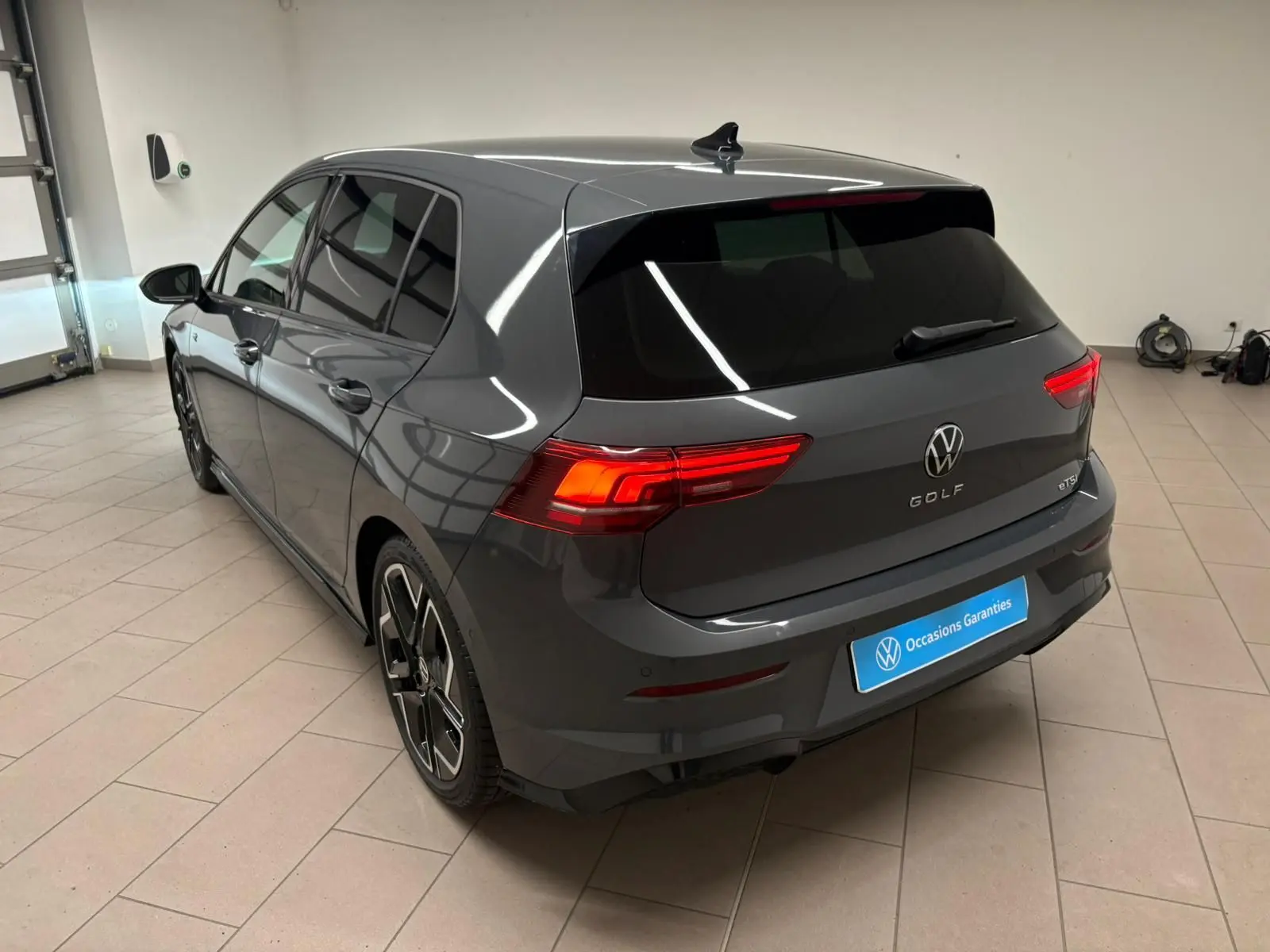 Vue 3/4 arrière d'une Volkswagen Golf 1.5 eTSI gris dauphin avec feux arrière allumés en intérieur showroom.