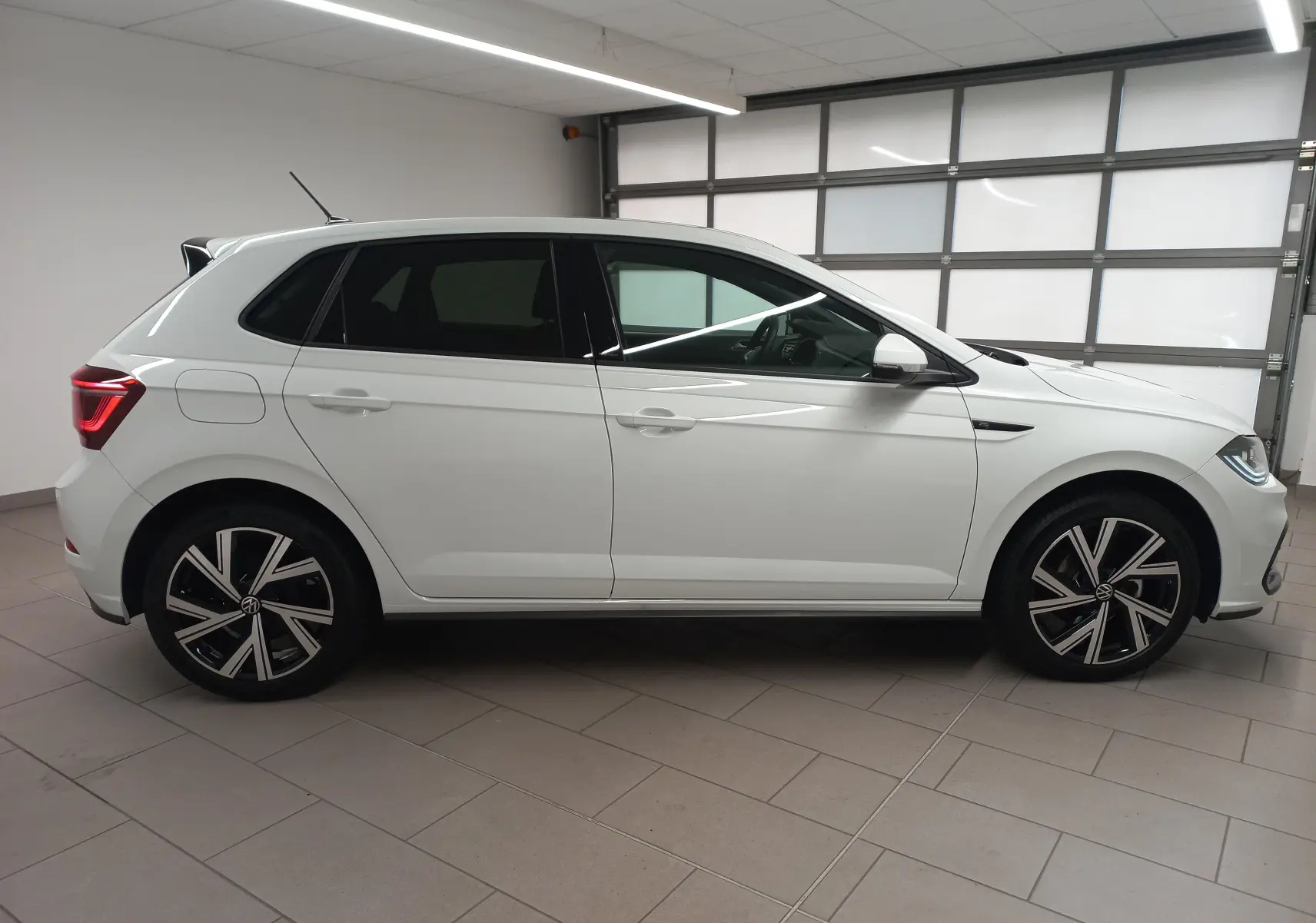 Vue de profil côté gauche d'une Volkswagen Polo blanche avec vitres arrière surteintées et jantes R-Line noires et argentées.
