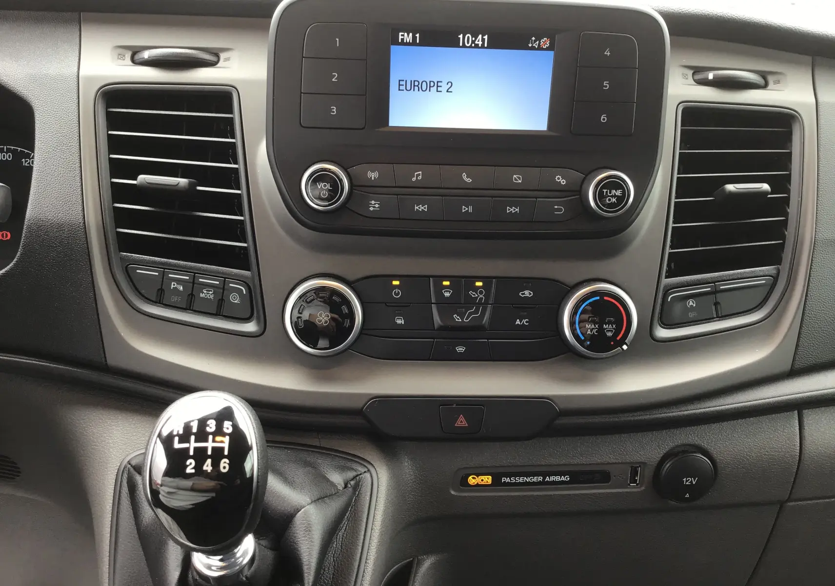 Tableau de bord du Ford Transit Fourgon 2023 avec écran radio, commandes climatisation et levier de vitesse manuel en vue frontale.