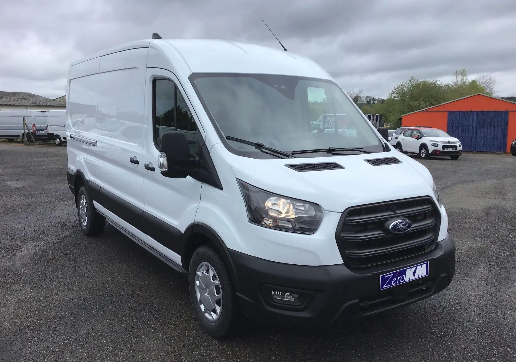 Fourgon Ford Transit blanc vu en 3/4 avant droit, avec calandre noire et feux allumés sur un parking extérieur.