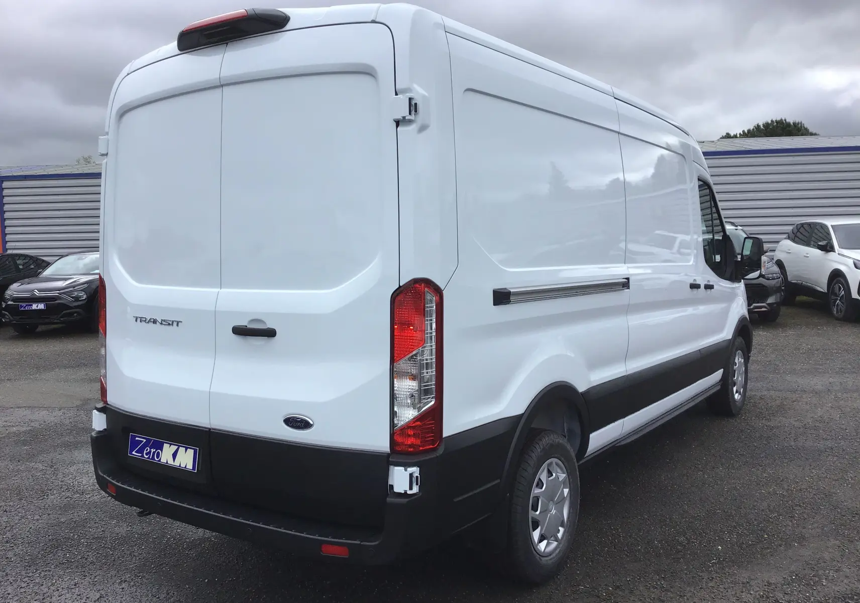Vue 3/4 arrière droite d'un Ford Transit Fourgon blanc 2023 avec hayon arrière fermé et protections noires.