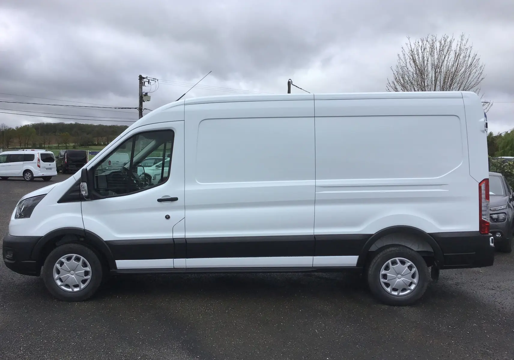 Vue de profil côté gauche d'un fourgon Ford Transit blanc 350 L3H2 avec bande noire basse sur un parking extérieur.