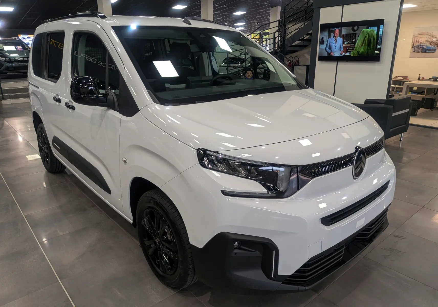 Vue 3/4 avant d'un Citroën Berlingo blanc Glacier avec jantes noires dans un showroom moderne.