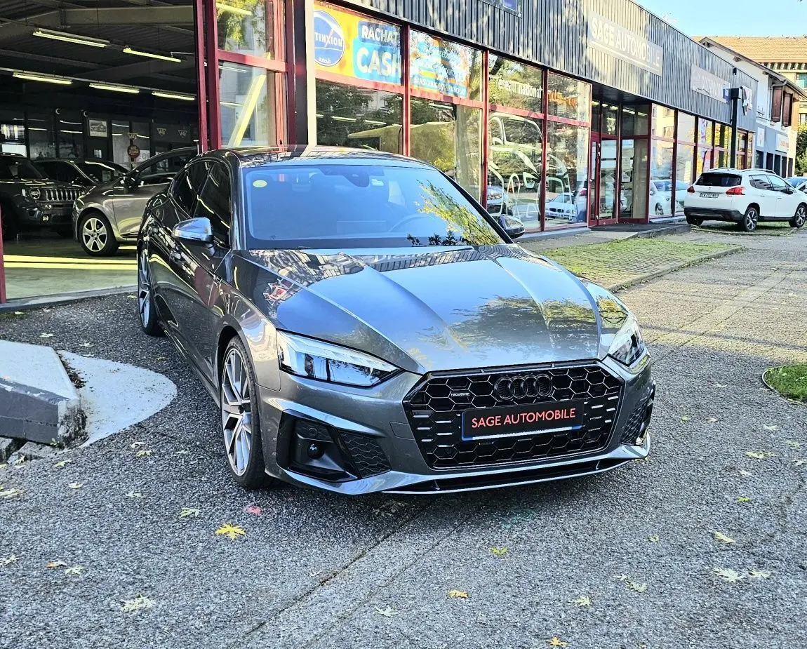 Audi A5 Sportback gris Daytona vue 3/4 avant, avec calandre noire hexagonale et jantes alliage visibles devant un garage.