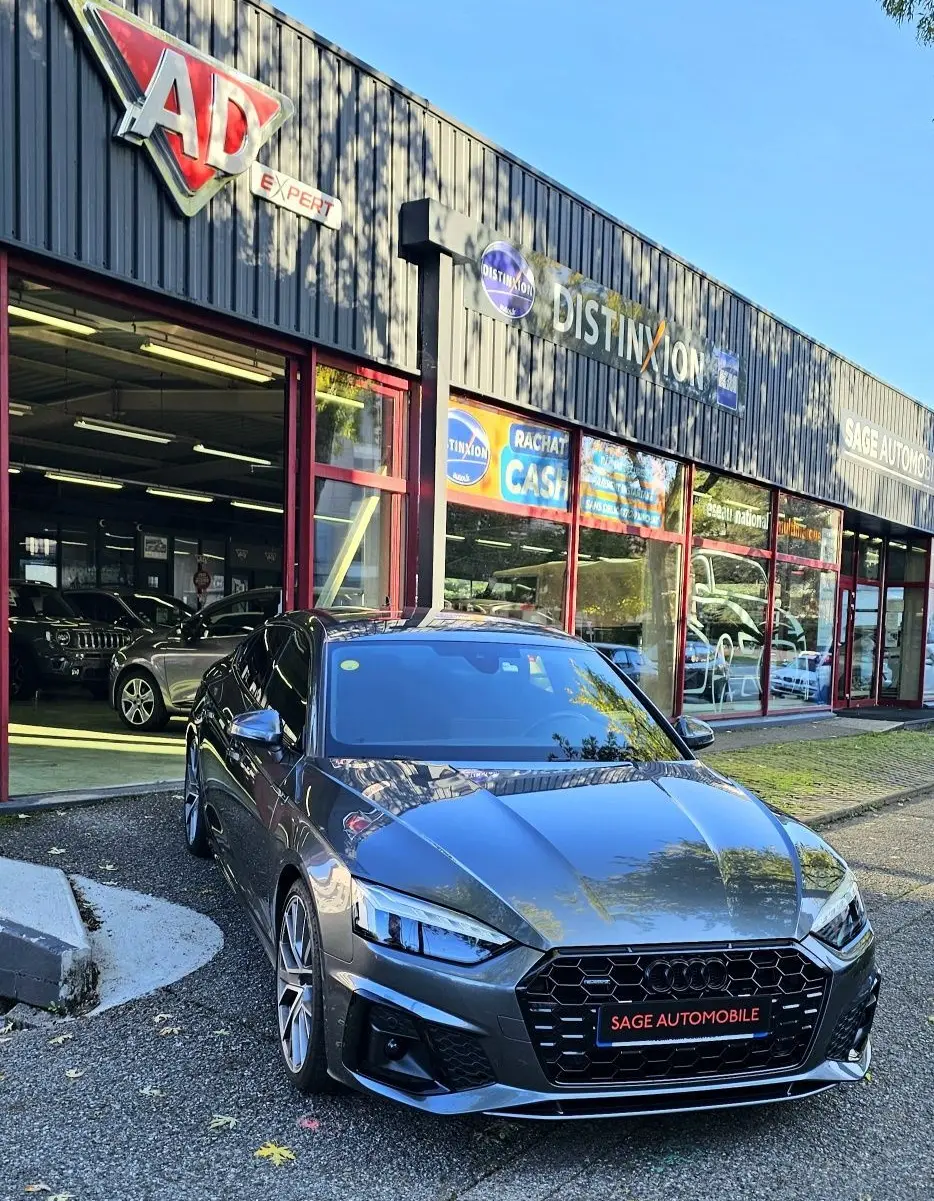 Audi A5 Sportback gris Daytona vue en 3/4 avant droit devant un garage avec enseignes visibles.