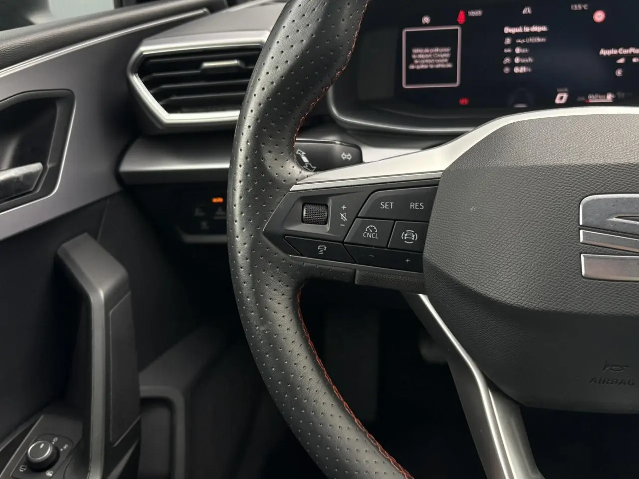 Gros plan sur le volant en cuir noir perforé avec surpiqûres rouges du SEAT Leon 2025, tableau de bord numérique en arrière-plan.