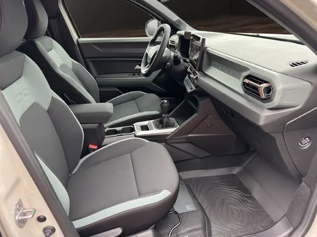 Intérieur avant droit du Dacia Duster 2025 gris clair, sièges noirs avec surpiqûres, tableau de bord moderne et volant simili cuir.