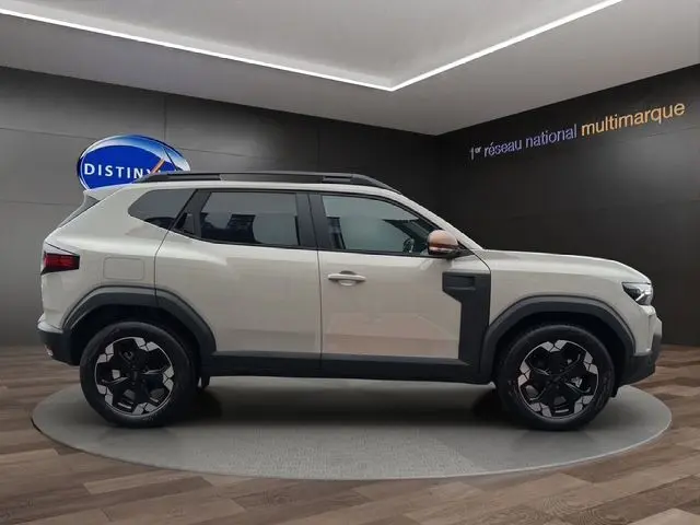 Profil latéral droit d'un Dacia Duster 2025 beige avec jantes alu noires et barres de toit en showroom.
