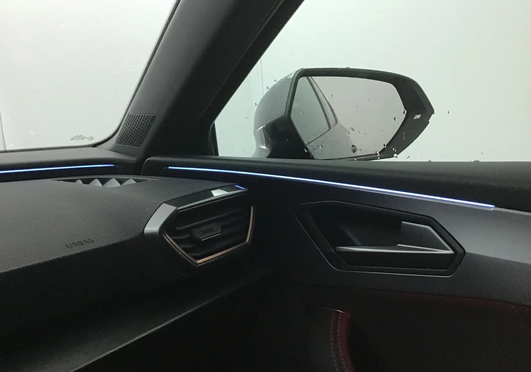 Intérieur côté passager d'une CUPRA Leon noire 2023, avec éclairage d'ambiance bleu et rétroviseur extérieur visible.