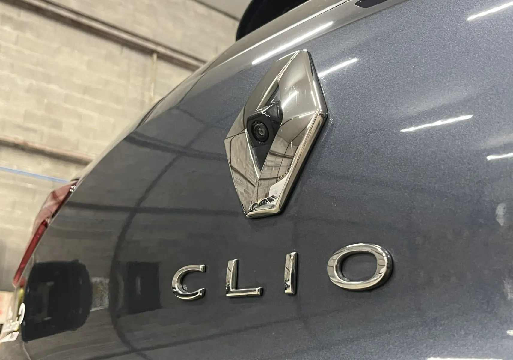 Gros plan sur le logo Renault et l’inscription Clio à l’arrière d’une Renault Clio V gris titanium dans un garage.