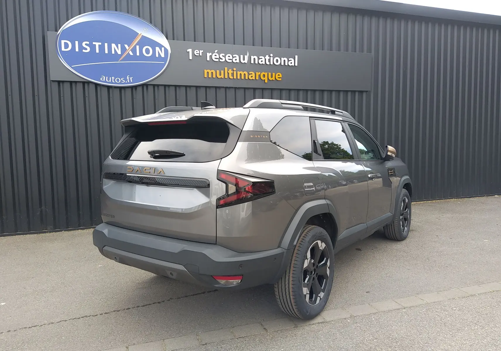 Vue 3/4 arrière droite du Dacia Bigster gris schiste avec jantes noires et logo doré sur un parking extérieur.