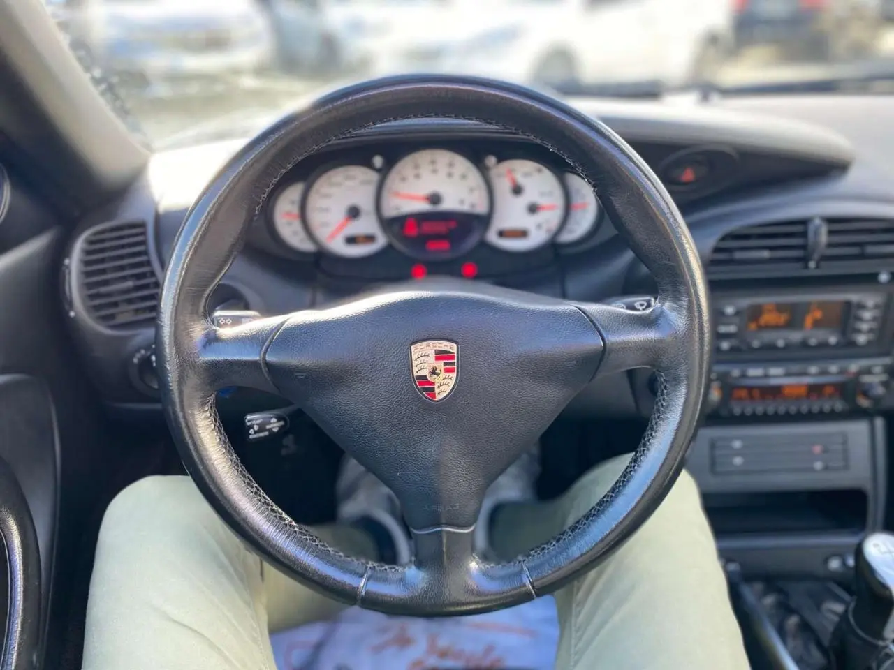 Vue intérieure centrée sur le volant en cuir noir avec logo Porsche, tableau de bord blanc et levier de vitesse manuel visible.