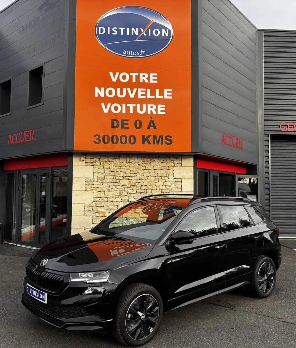 SKODA KAROQ noir vue 3/4 avant droit, garée devant un bâtiment avec panneau orange Distinxion.