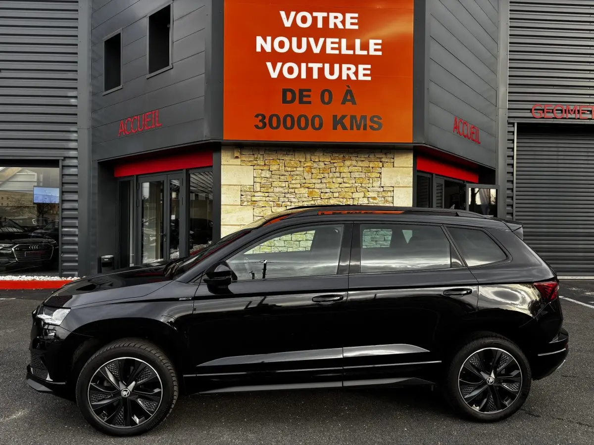 Profil côté gauche d'un SUV SKODA KAROQ noir 2025 garé devant un bâtiment avec panneau orange "Votre nouvelle voiture".