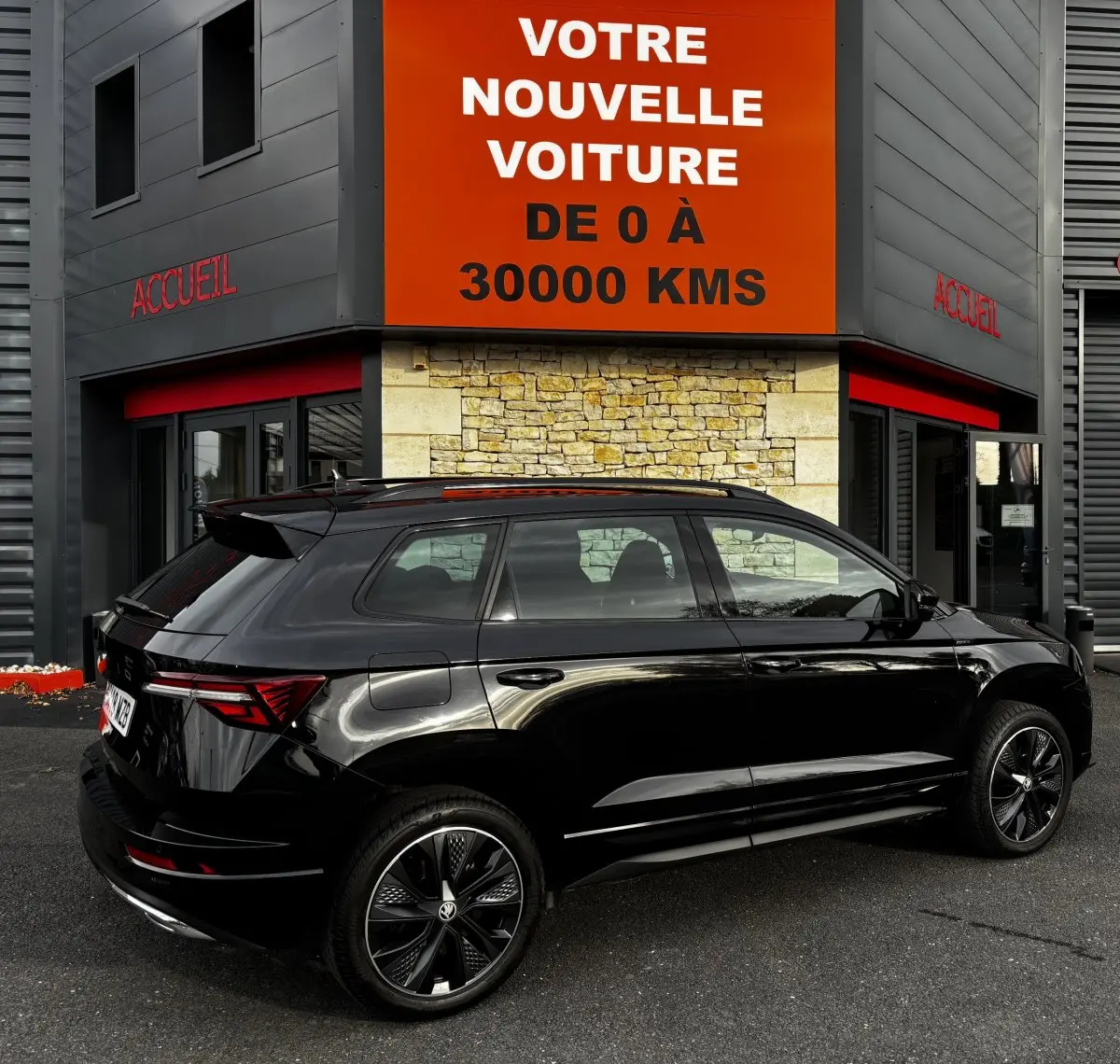 Vue 3/4 arrière droite d'un SUV SKODA KAROQ noir brillant garé devant un bâtiment avec une enseigne orange.