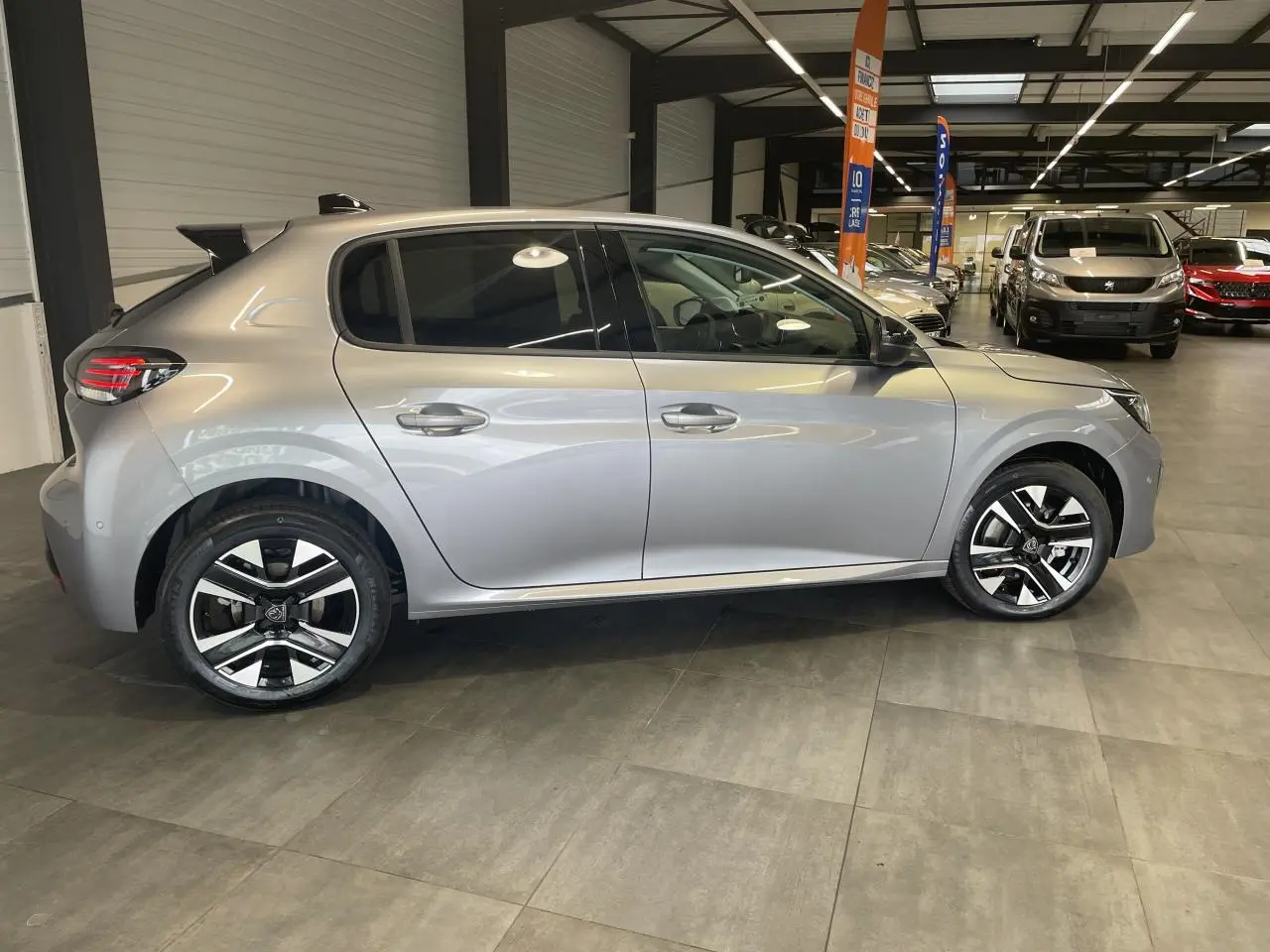 Profil côté gauche d'une Peugeot 208 1.2i Hybrid gris Artense avec jantes alliage bi-tons en showroom.