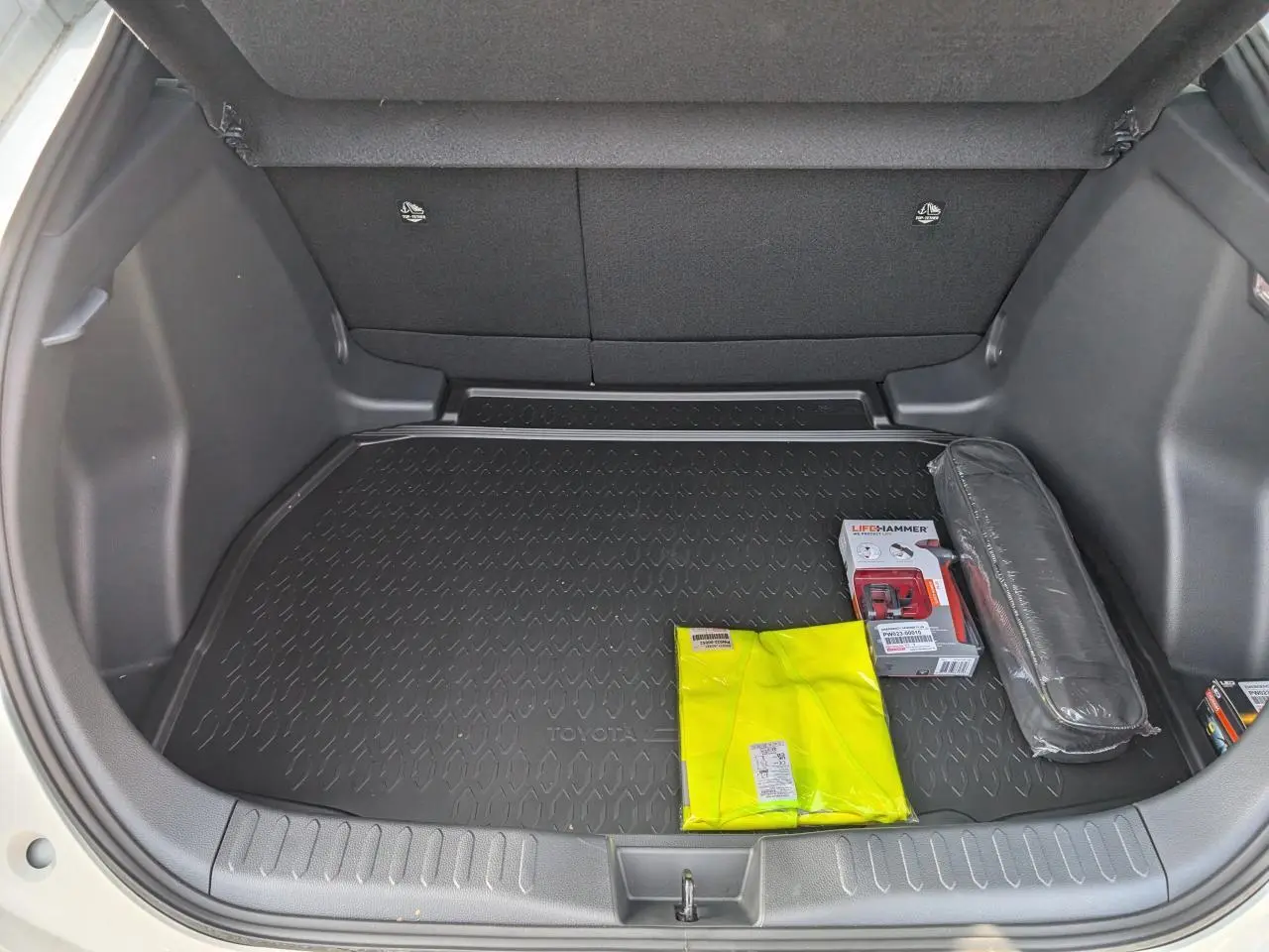 Coffre ouvert d’un Toyota C-HR blanc, vue de dessus montrant tapis noir et kit de sécurité jaune et noir.