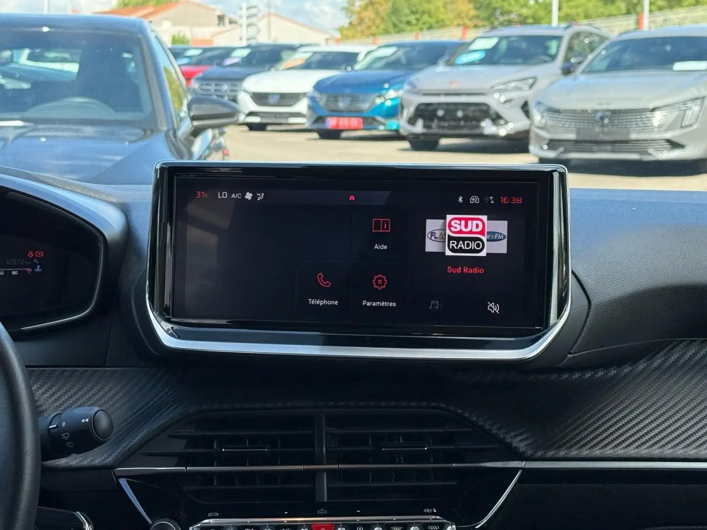 Vue intérieure centrée sur l'écran tactile du tableau de bord de la Peugeot 208 gris Artense, avec vue sur le pare-brise et voitures au loin.