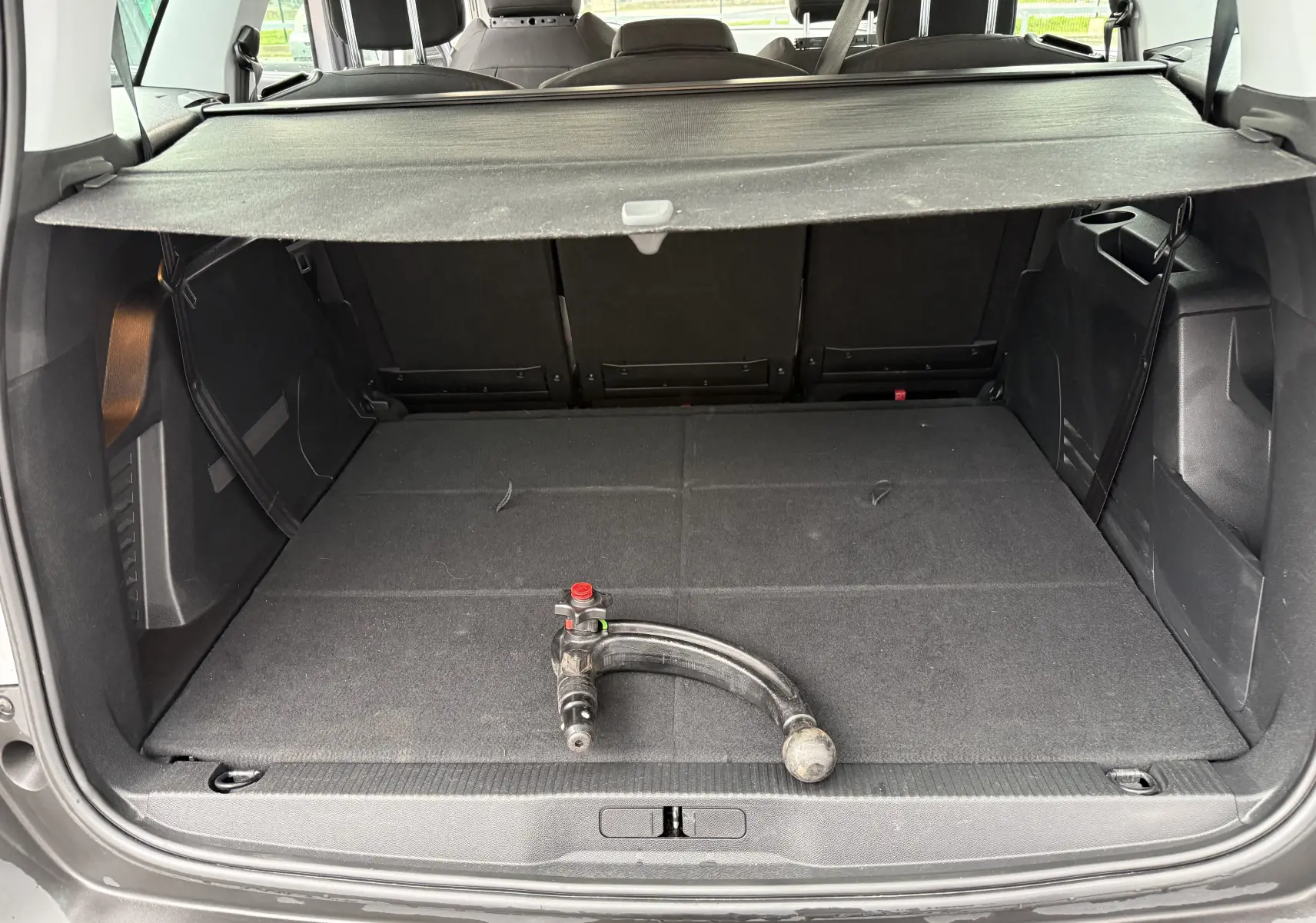 Coffre ouvert d'un Peugeot 5008 gris Artense, vue arrière montrant la plage arrière et une barre d'attelage posée au sol.