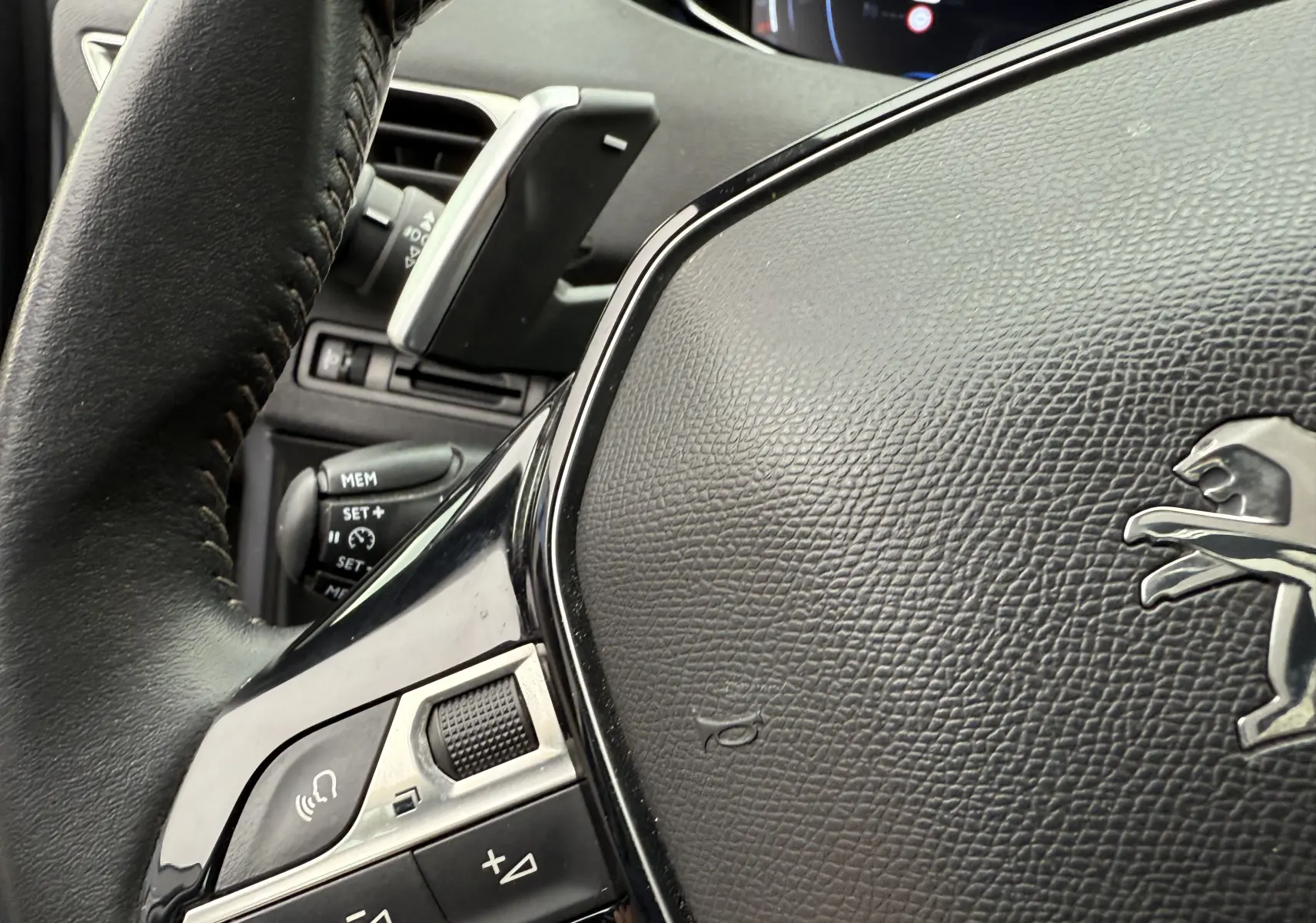 Gros plan sur le volant en cuir noir du Peugeot 5008 gris Artense, avec commandes intégrées et logo lion visible.