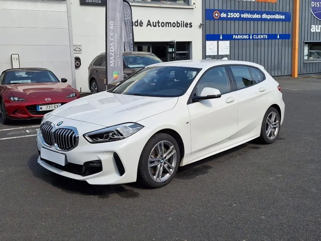 BMW Série 1 120i M Sport blanc vue 3/4 avant droit, avec jantes aluminium et calandre imposante.