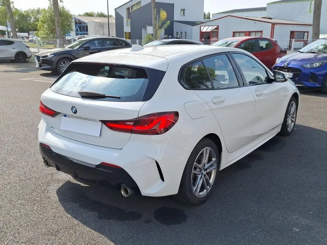 BMW Série 1 blanche vue en 3/4 arrière droit, avec jantes aluminium et feux arrière LED distinctifs.