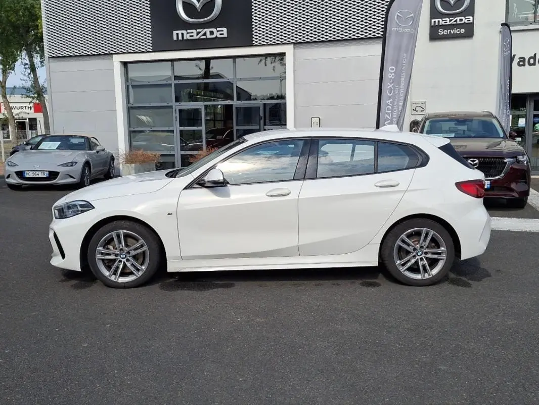 Profil côté gauche d'une BMW Série 1 blanche 120i M Sport 2021, avec jantes aluminium et lignes sportives visibles.