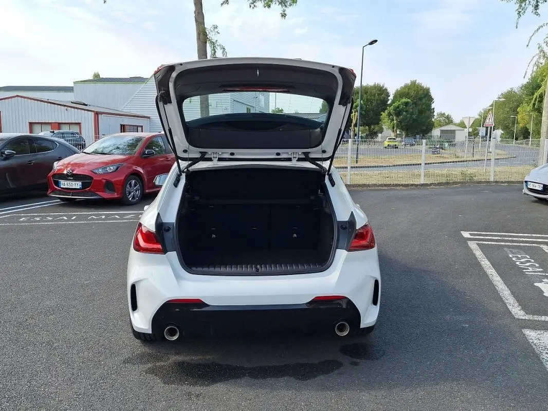 Vue arrière d'une BMW Série 1 blanche 120i M Sport 2021 avec coffre ouvert sur un parking extérieur.