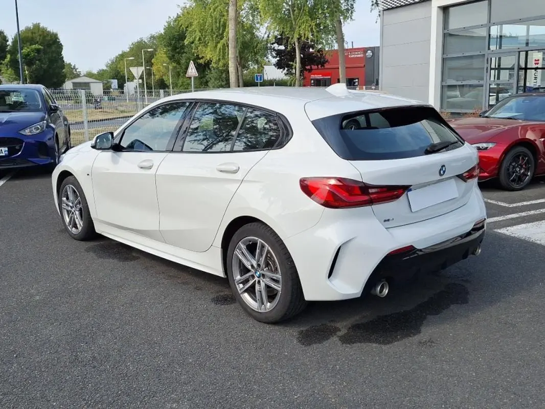 BMW Série 1 120i M Sport blanc vue 3/4 arrière droit, avec jantes aluminium et double sortie d’échappement chromée.