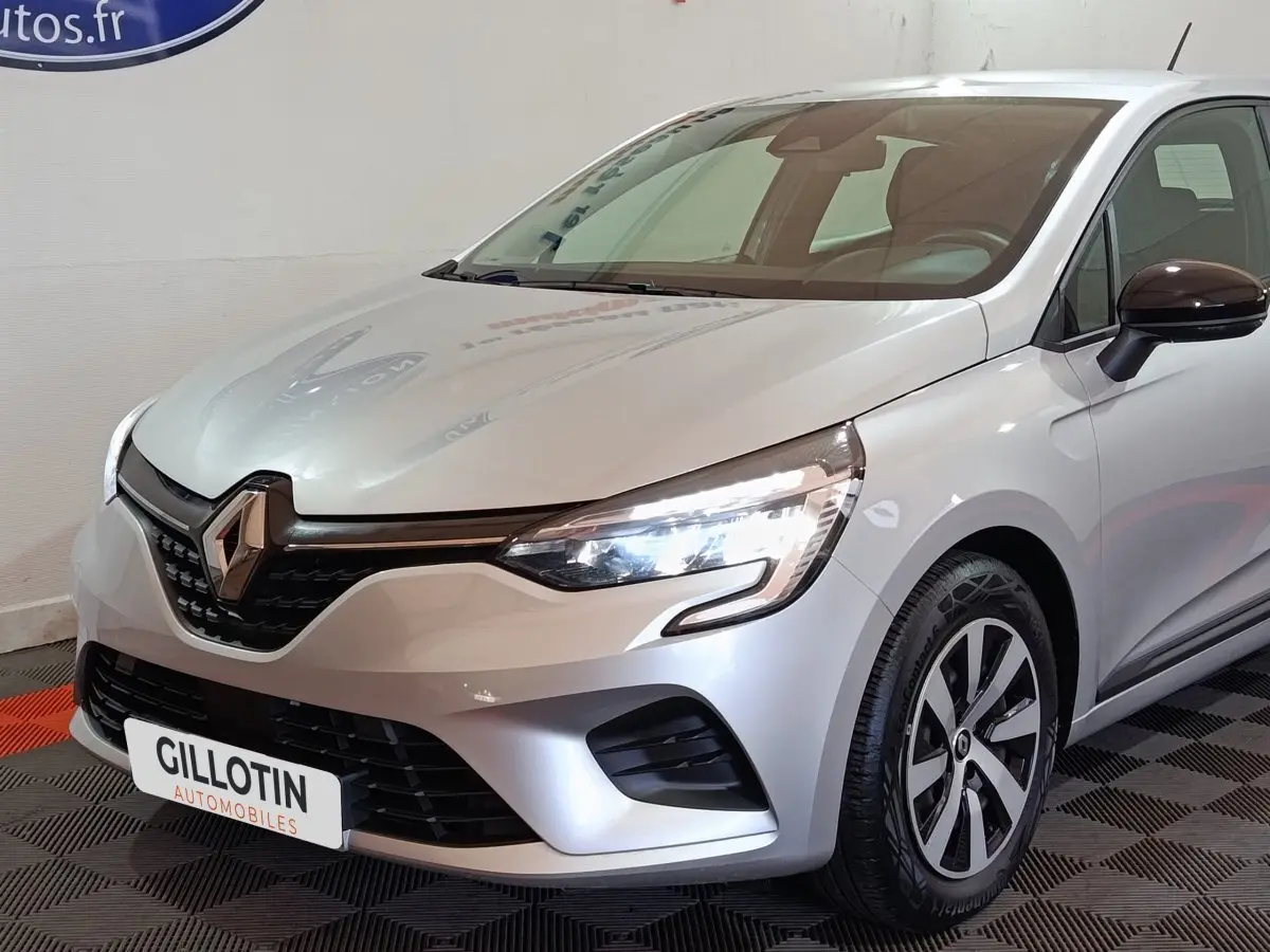 Vue 3/4 avant droite d'une Renault Clio gris clair 2023 avec phares LED allumés et jantes distinctives.