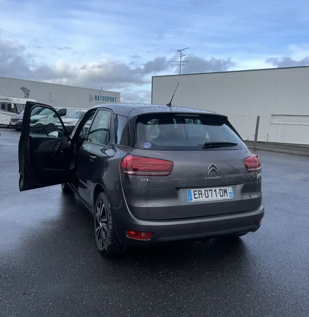 Vue 3/4 arrière gauche d'une Citroën C4 Picasso marron 2017 avec porte avant gauche ouverte sur parking humide.