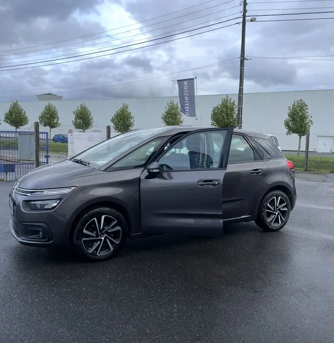 Vue de profil côté gauche d'une Citroën C4 Picasso marron avec porte avant ouverte et jantes alu sur sol humide.