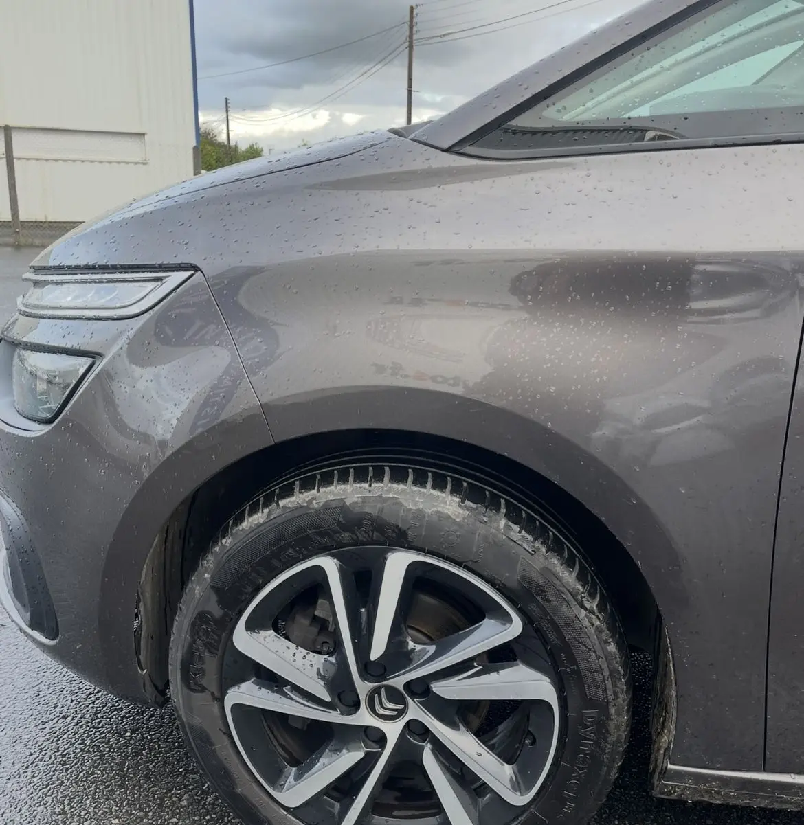 Gros plan sur la roue avant gauche et l'aile d'une Citroën C4 Picasso marron sous la pluie avec jante alu bicolore.