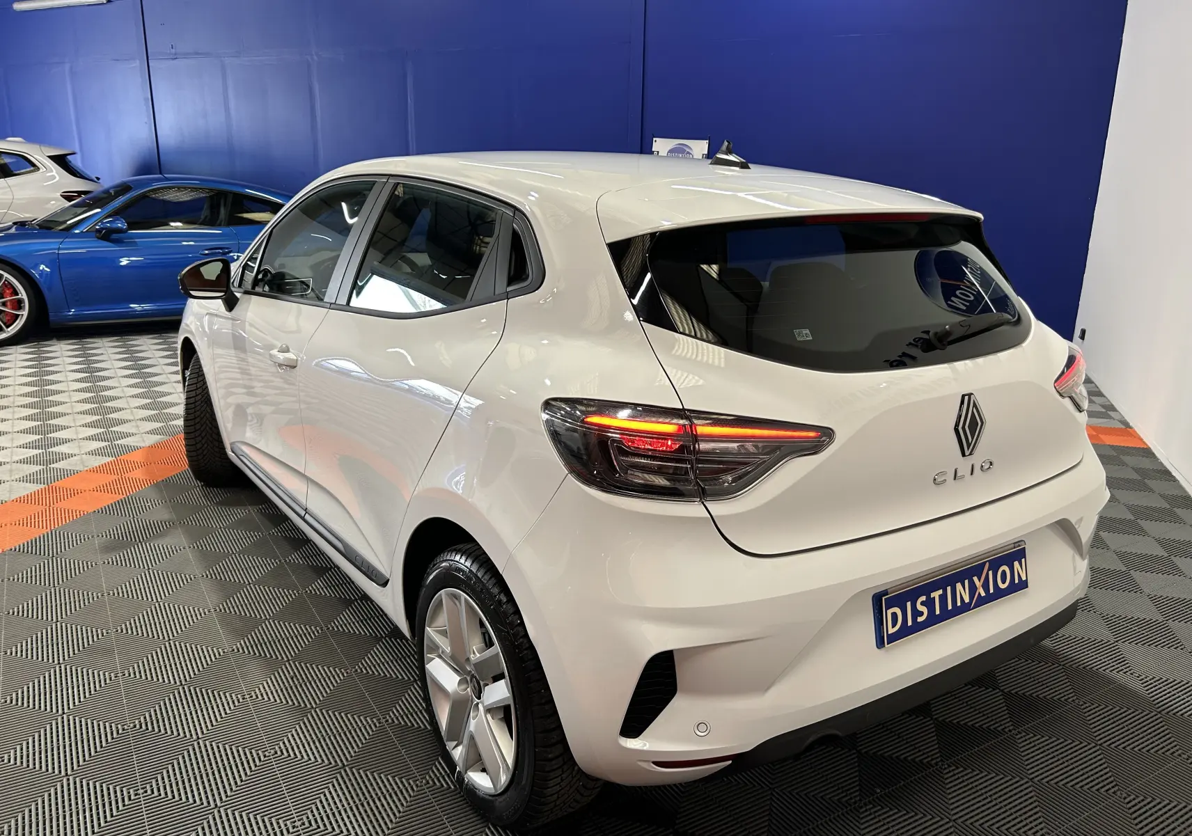 Vue 3/4 arrière droite d'une Renault Clio V blanche avec feux LED allumés, garée sur un sol à damier en intérieur.