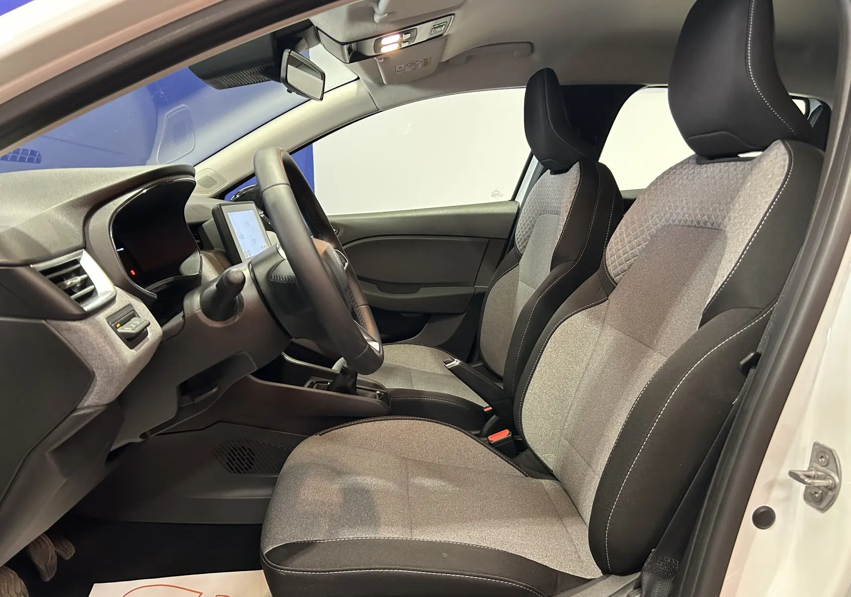 Vue intérieure côté conducteur de la Renault Clio V blanche, sièges tissu gris avec surpiqûres blanches et volant cuir.