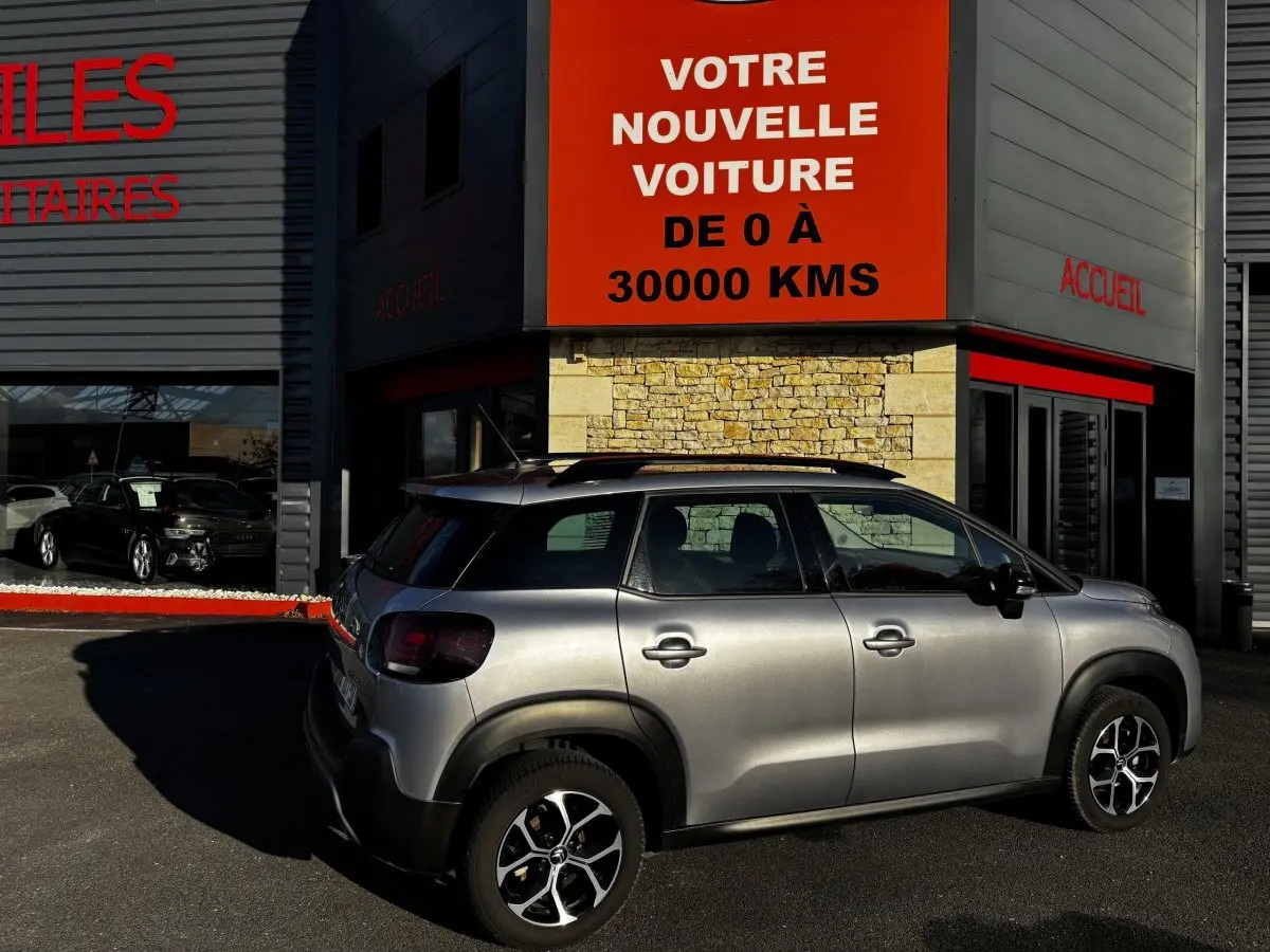 Citroën C3 Aircross gris clair vue de profil côté droit, stationnée devant un bâtiment avec panneau orange.