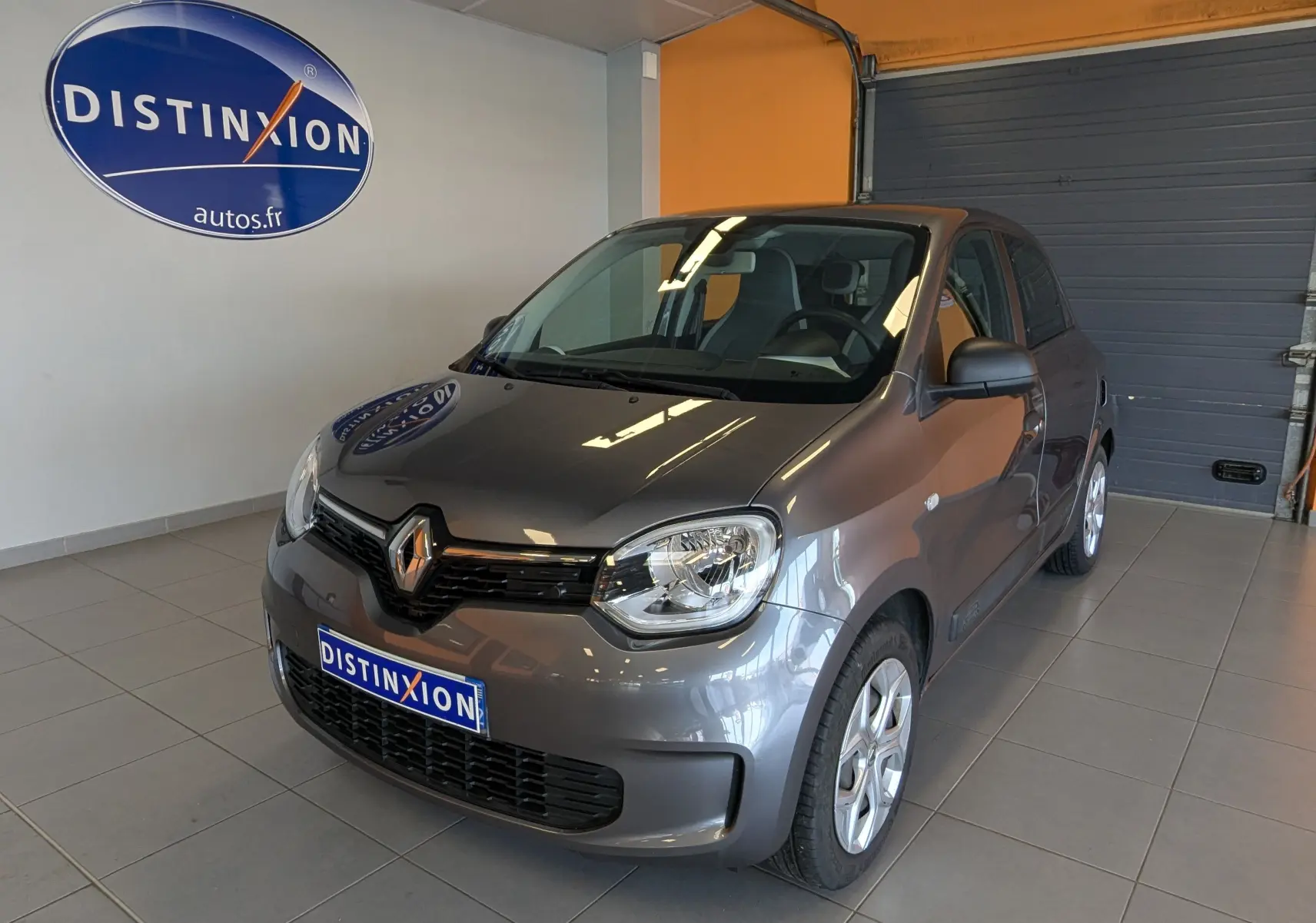 Renault Twingo III gris cassiopée vue 3/4 avant droit en intérieur avec logo Distinxion en arrière-plan.