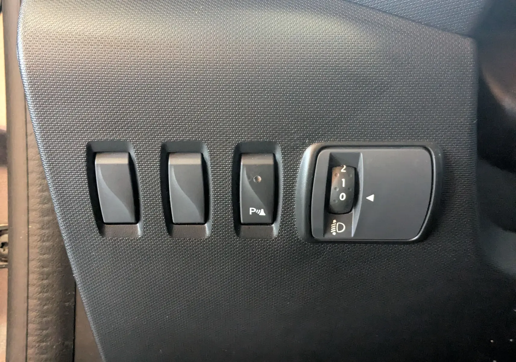 Gros plan sur les commandes de réglage des phares et aides à la conduite dans une Renault Twingo III gris cassiopée, côté conducteur.