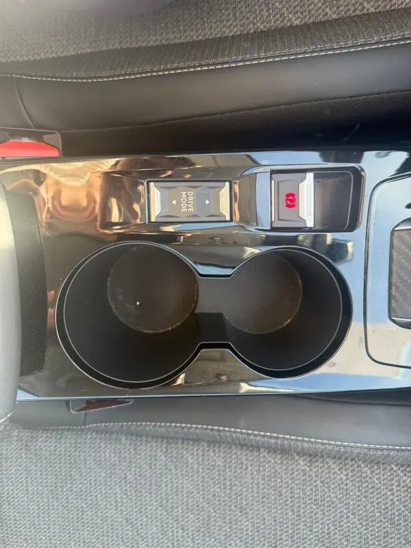 Gros plan sur la console centrale noire de la Peugeot 208 Hybrid 110 e-DCS6 Allure 2024, avec porte-gobelets et bouton Drive Mode.
