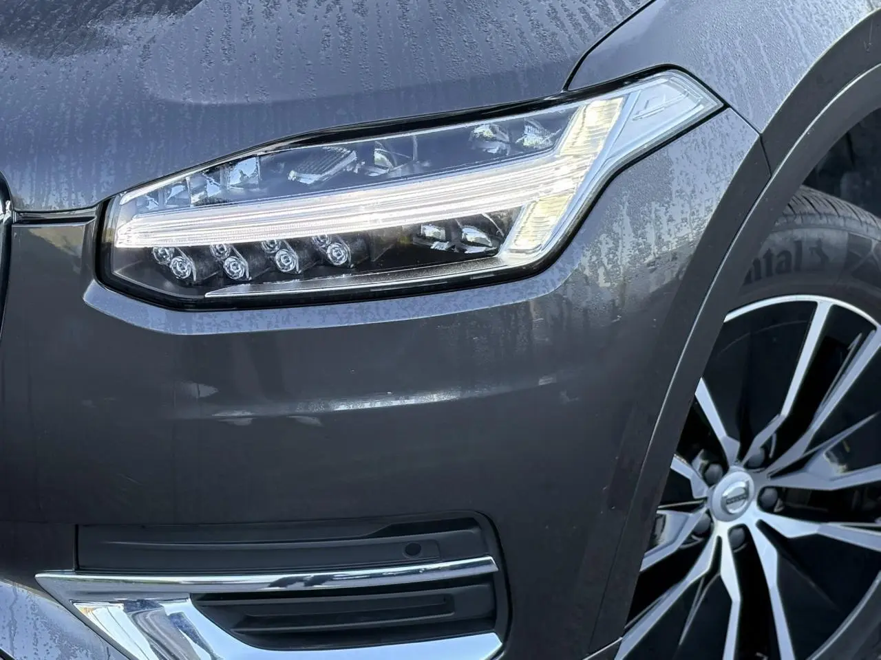 Gros plan sur le phare avant gauche et la jante alliage 19" noir diamant du Volvo XC90 gris Platinium 2024.