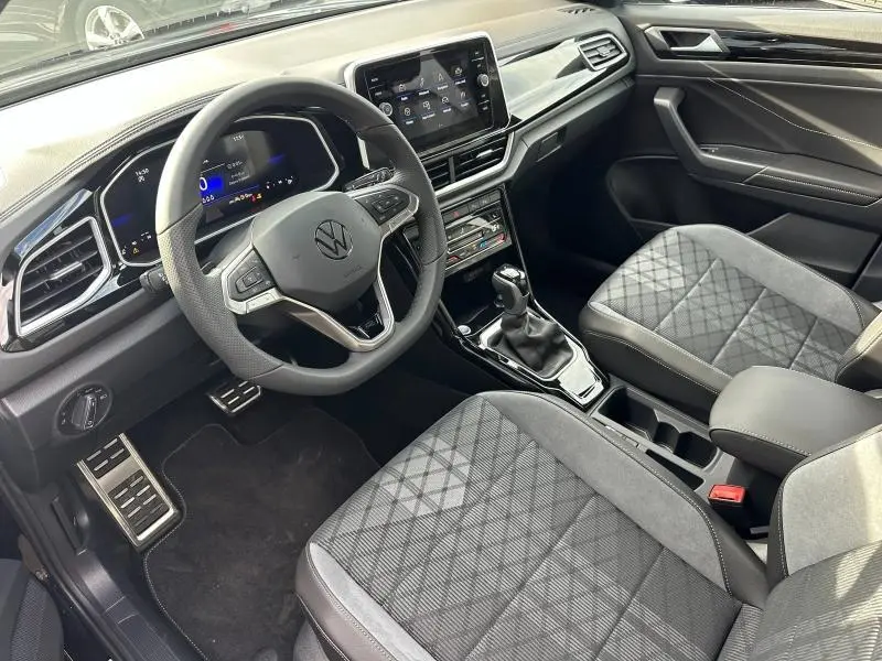 Vue intérieure côté conducteur du Volkswagen T-Roc 2025, avec volant multifonction, sièges gris à motifs et console centrale moderne.