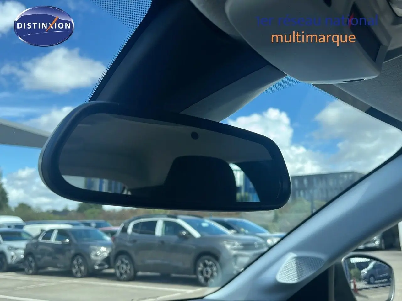 Rétroviseur intérieur reflétant le profil droit d'une Opel Corsa gris kristal métal stationnée en extérieur sous un ciel partiellement nuageux.