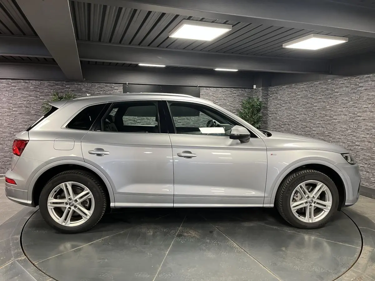 Audi Q5 gris en vue profil côté gauche dans un showroom, avec jantes alliage et badge S line visible sur l'aile avant.
