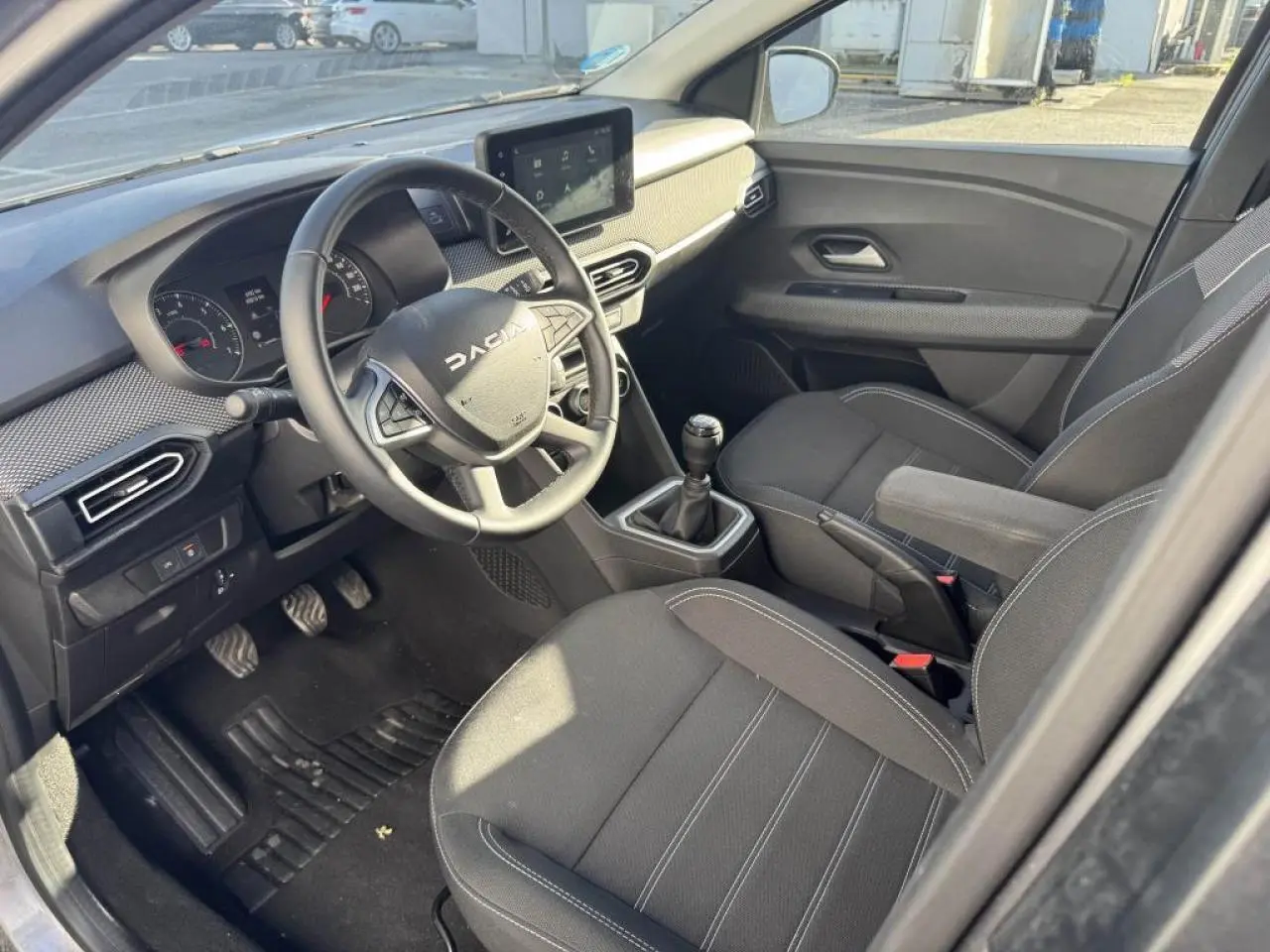 Vue intérieure côté conducteur du Dacia Jogger 2025, montrant le volant, la console centrale et les sièges noirs.