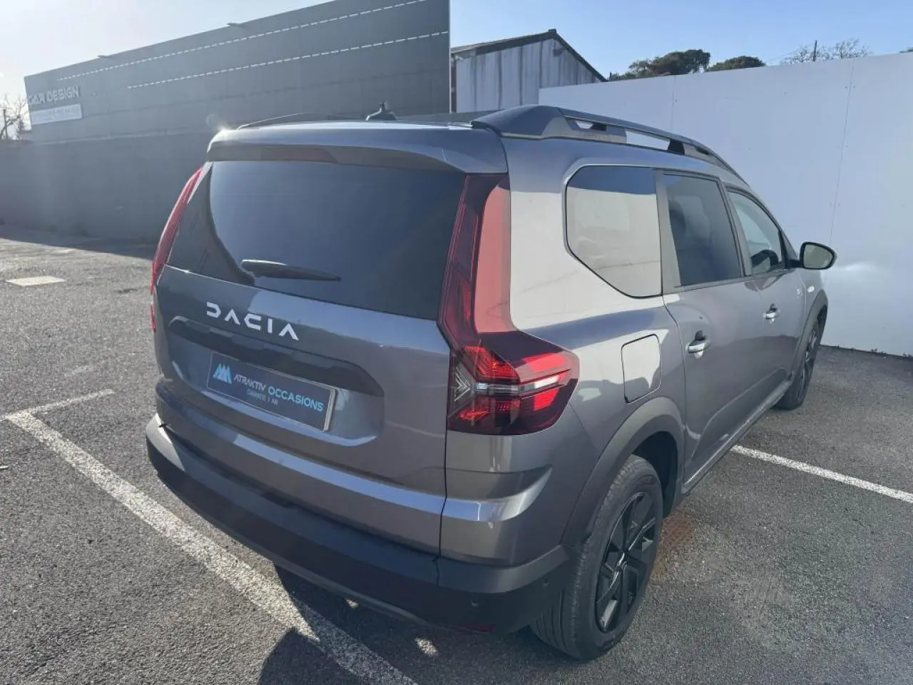 Vue 3/4 arrière droit d'un Dacia Jogger gris schiste métal avec barres de toit et feux arrière LED distinctifs.