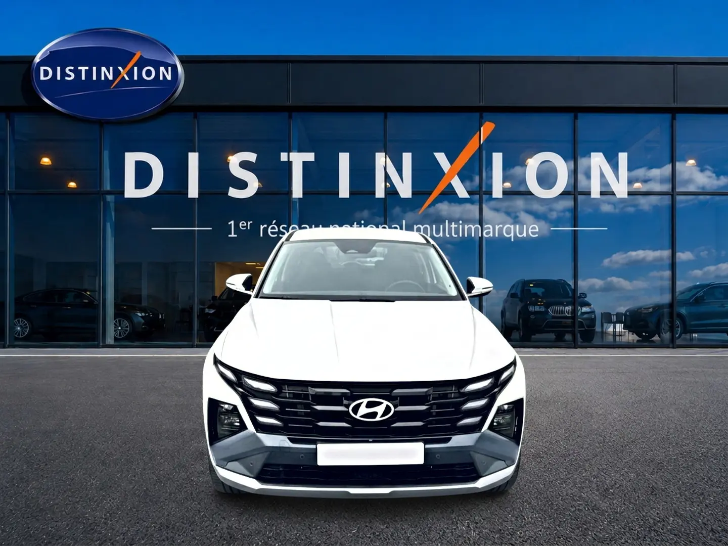 Vue frontale d’un Hyundai Tucson blanc 2026 avec calandre noire distinctive, devant une vitrine de concession multimarque.