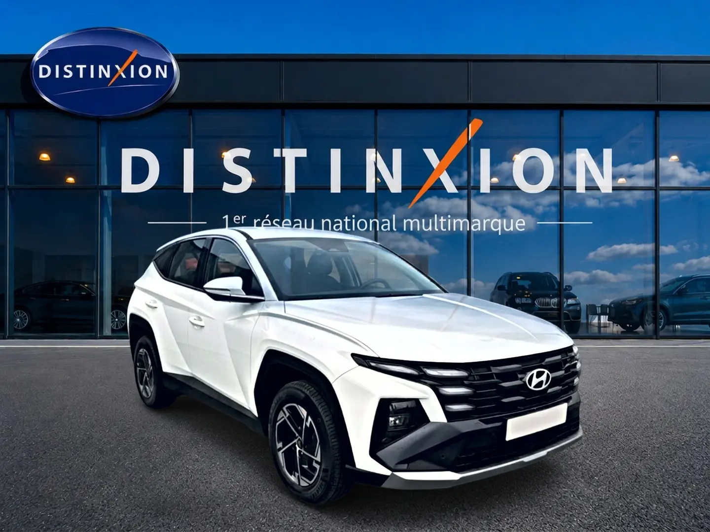 Vue 3/4 avant gauche d’un Hyundai Tucson blanc 2026 avec jantes alliage et calandre noire devant un bâtiment vitré Distinxion.